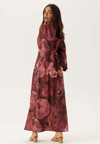 Maxiklänning i genomskinligt vinrött tyg med stora blommönster. Har långa ärmar och en rynkad ryggdesign. Bärs med nudefärgade klackskor.