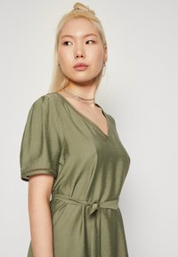 Maslinasto zelena haljina s V-izrezom, kratkim napuhanim rukavima, detaljem vezanja u struku i suptilnom teksturom tkanine. Srebrni nakit upotpunjuje izgled.