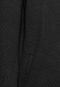 Tessuto nero texturizzato con un motivo a diamante, con una cucitura e dettagli della tasca visibili. La superficie appare morbida e leggermente in rilievo.