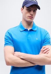 Νεαρός άνδρας με σταυρωμένα τα χέρια, φορώντας μπλε πόλο και σκούφο ναυτικού μπλε, μπροστά σε απλό ανοιχτό φόντο.