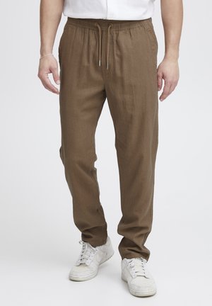 Mand iført brune bukser med snøre og hvide sneakers, stående mod en ensfarvet lys baggrund.