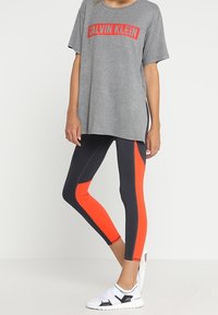 Szara, oversize'owa koszulka z czerwoną grafiką "CALVIN KLEIN", w połączeniu z czarnymi legginsami z pomarańczowymi panelami po bokach oraz białymi butami sportowymi.