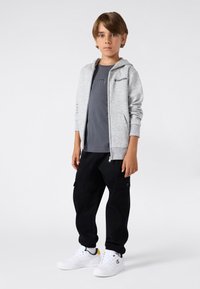 Niño de pie con sudadera gris con cremallera, camiseta gris oscuro, pantalones cargo negros y zapatillas blancas sobre un fondo liso.
