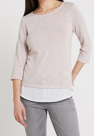 Kobieta ubrana w jasnoróżową bluzkę z rękawami do łokcia, nałożoną na białą koszulę, w połączeniu z jasnoszarymi dżinsami, stojąca z jedną ręką w kieszeni.