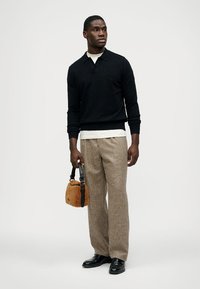 Pulôver preto de malha, calças bege de padrão xadrez e sapatos pretos. Asegura uma pequena bolsa de pele castanha com uma alça. Design simples e estruturado.