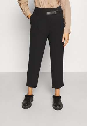Vrouw draagt zwarte high-waist cropped broek met zijzakken, een zwarte riem, een beige ingestopte blouse en zwarte bontgevoerde instappers.