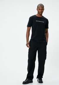 Sort bomuld t-shirt med korte ærmer og hvid tekst trykt over brystet, parret med sorte cargo-bukser med sidelommer.