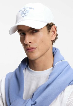 Junger Mann trägt weiße Tenniscap mit blauem Logo, weißes Hemd und hellblauen Pullover über den Schultern vor einfachem Hintergrund.