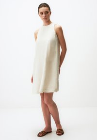 Ärmelloses, knielanges Kleid aus leichtem beige Leinen mit lässiger Passform, das durch klare Linien und eine subtile Textur besticht. Trägt dazu braune Sandalen.