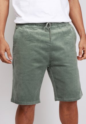 Uomo che indossa pantaloncini verdi di velluto a coste con coulisse e una maglietta bianca, in piedi su uno sfondo semplice.