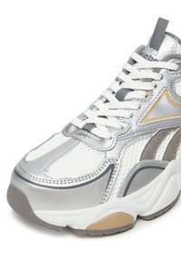 Sudraba un baltas sporta kedas ar baltām auklām, acs paneļiem un bēšiem akcentiem uz zoles un sāna pie papēža.