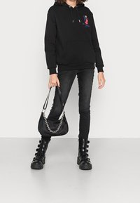 Černá mikina s kapucí a přední kapsou, ozdobená barevným vyšíváním, v kombinaci s černými skinny džíny a robustními černými botami se sponami.