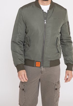 Homme portant une veste bomber vert olive avec poches avant et fermeture éclair, associée à un short cargo marron sur fond blanc.
