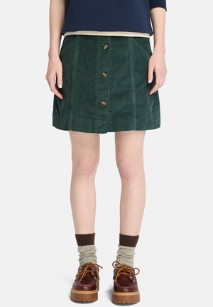 Groene corduroy rok met een knoopsluiting, voorzakken en een gestructureerde zoom. Gecombineerd met een paar bruine schoenen en getextureerde sokken.