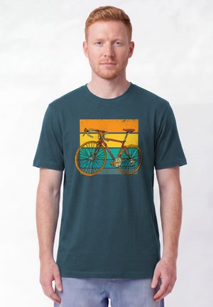 Mann mit kurzen roten Haaren trägt ein blaues T-Shirt mit einer Grafik eines Vintage-Rennrads vor einem Hintergrund mit orange-, gelb- und türkisfarbenen Streifen.