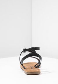Musta nahkasandaali, jossa on teksturoitu hihnadesign, nilkkaluukku ja beige keinonahkainen pohja. Litteä pohja, jossa on minimaalinen kuviointi.