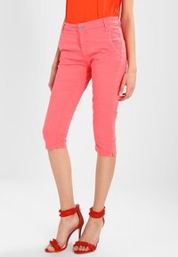 Růžové capri kalhoty z bavlny se slim fit střihem, s předními kapsami a zapínáním na knoflík. Na nohou má červené sandály na podpatku.