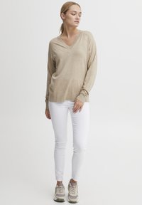 Beige V-kivágású pulóver hosszú ujjakkal, fehér slim-fit nadrággal párosítva. A modellen világosszürke sportcipő van, sötétebb szürke részletekkel.