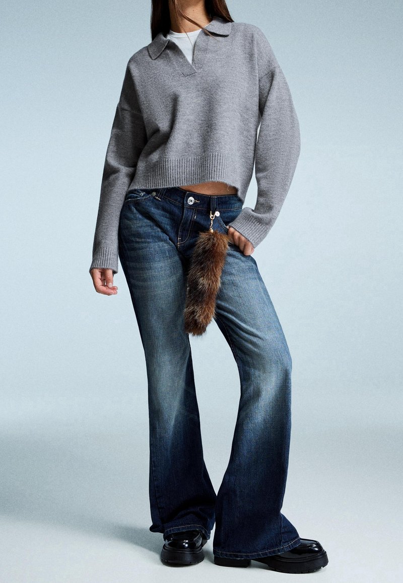 Personne portant un pull gris à manches longues, un jean évasé foncé avec un porte-clés en fourrure et des chaussures noires sur un fond clair uni.