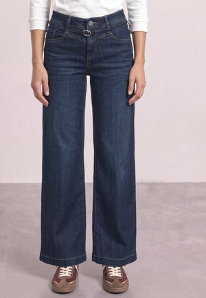 Person iført mørkeblå jeans med vide ben, hvid top og brune sneakers, som står på en grå gulv.