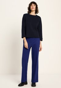 Frau, die in einem schwarzen Langarmoberteil, blauen weit geschnittenen Hosen und schwarzen Stiefeletten vor einem beigen Hintergrund steht.