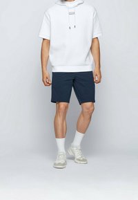 Bílá mikina s kapucí a krátkým rukávem, doplněná modrými šortkami. Model má na sobě bílé ponožky a světle šedé sportovní boty.