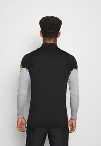 Schwarzes Sportshirt mit langen grauen Ärmeln, die eine gerippte Textur und neon-grüne Akzente an der Schulter aufweisen. Schlankes Design.