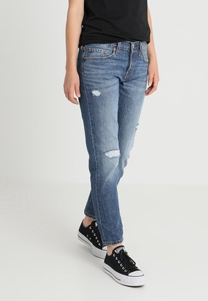 Persoon draagt lichtblauwe gescheurde spijkerbroek, zwarte sneakers en een zwart T-shirt, staand tegen een effen lichte achtergrond.