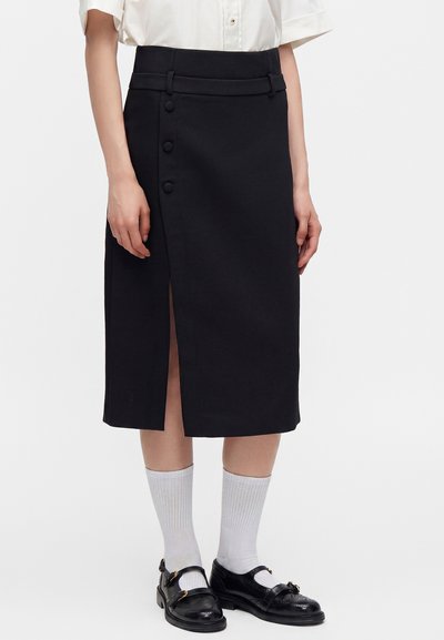 Jupe noire mi-longue avec une fente devant et trois boutons, portée avec des chaussettes blanches, des chaussures noires à boucle et un chemisier blanc à manches courtes.