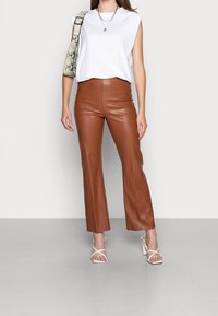 Vrouw in een mouwloze witte top, bruine leren broek, witte hoge hakken, en met een kleine schoudertas van slangenleerpatroon.