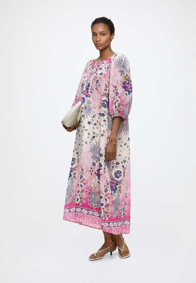 Mujer con un vestido largo y suelto de flores rosa y blanco con mangas tres cuartos, sosteniendo un clutch blanco y llevando tacones beige con tiras.