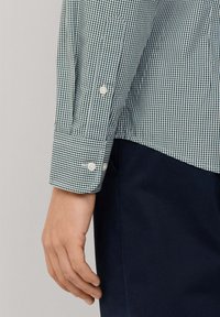 Camisa de cuadros verdes y blancos con puño abotonado. La tela es ligera y con textura. Pantalones azul marino parcialmente visibles en la parte inferior.