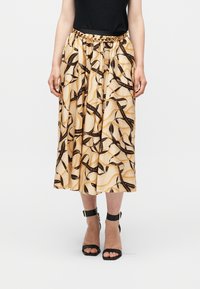 Μπεζ midi φούστα με μαύρο και χρυσό σχέδιο αλυσίδας και κορδέλας, συνδυασμένη με μαύρο αμάνικο τοπ, ζώνη με χρυσή αλυσίδα και μαύρα τακούνια με λουράκι στον αστράγαλο.