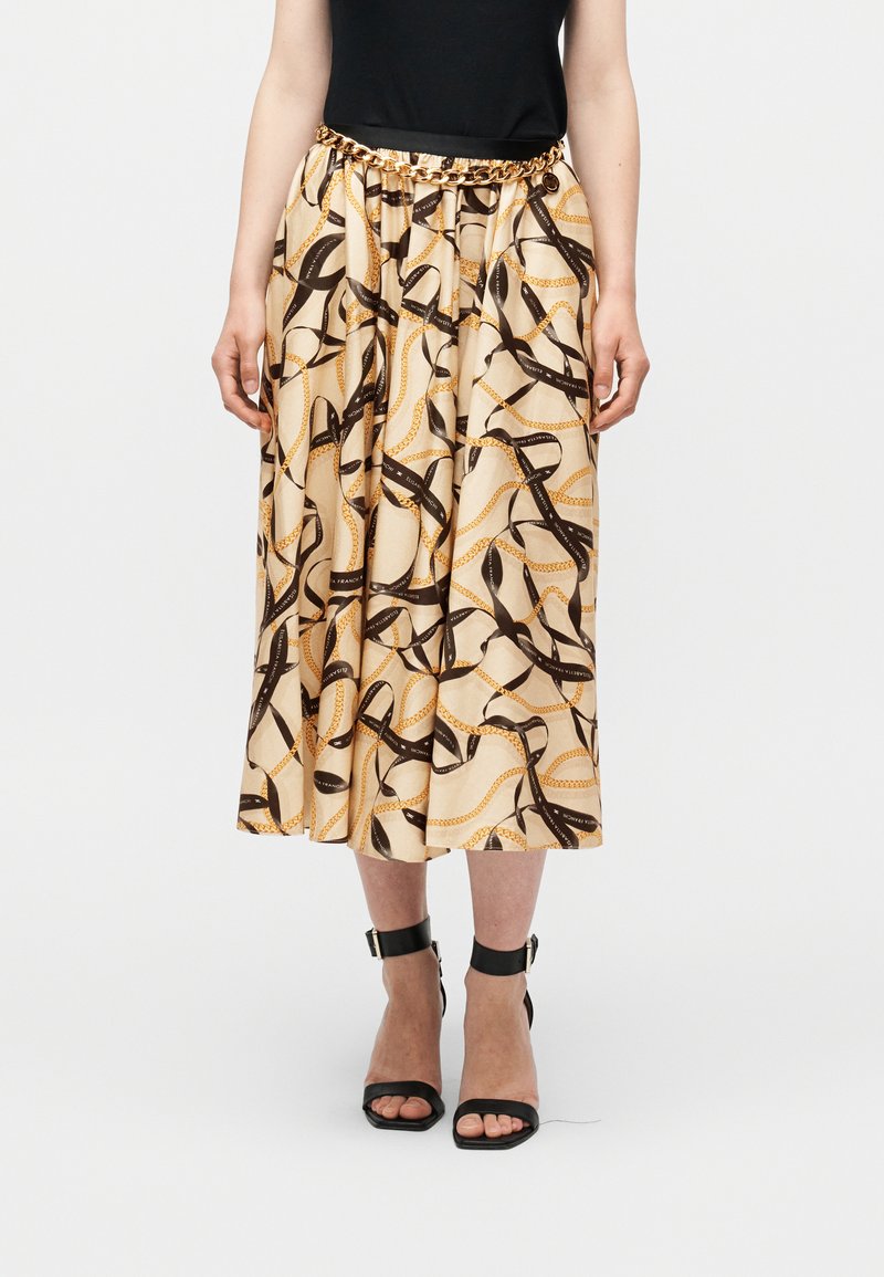 Μπεζ midi φούστα με μαύρο και χρυσό σχέδιο αλυσίδας και κορδέλας, συνδυασμένη με μαύρο αμάνικο τοπ, ζώνη με χρυσή αλυσίδα και μαύρα τακούνια με λουράκι στον αστράγαλο.