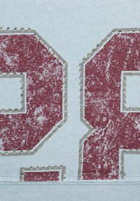 Vybledlý vínově červený číslo "24" obrysem z malých stříbrných hřebíčků na světle modré texturované látce.