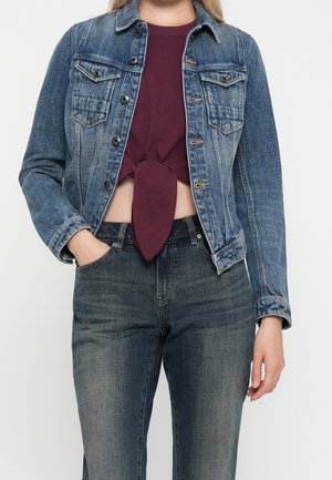 Osoba ubrana w niebieską kurtkę dżinsową nawiązującą na bordowy, zawiązany crop top i ciemne, wyblakłe dżinsy, z jedną ręką w kieszeni, stojąca na białym tle.