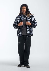 Faux pälsjacka i svart och blå kamouflage, med dragkedja framtill, kombinerad med en grå t-shirt och svarta vida byxor med grafik.