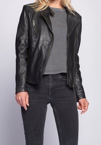 Žena oblečená v nerozepnuté černé kožené bundě přes šedé tričko a černé džíny, stojící na jednobarevném světlém pozadí.