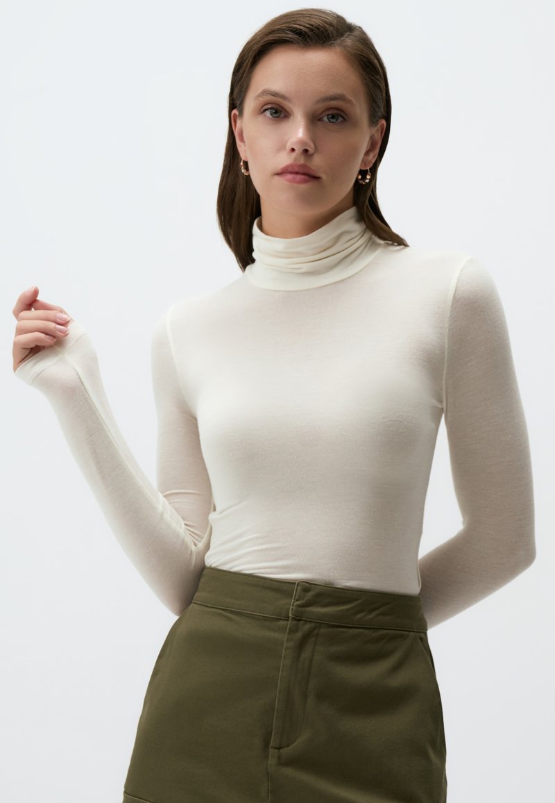 Maglietta a maniche lunghe color crema con colletto a dolcevita realizzata in tessuto leggero; design aderente con un leggero lucido e maniche lunghe; abbinata a pantaloni verde oliva.