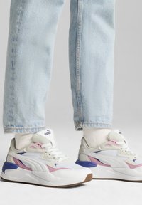 Weiße und graue Sneakers mit rosa und lila Akzenten, die eine strukturierte Sohle, auffällige Schnürsenkel und ein Logo auf der Zunge aufweisen. Getragen mit hellblauen Jeans.