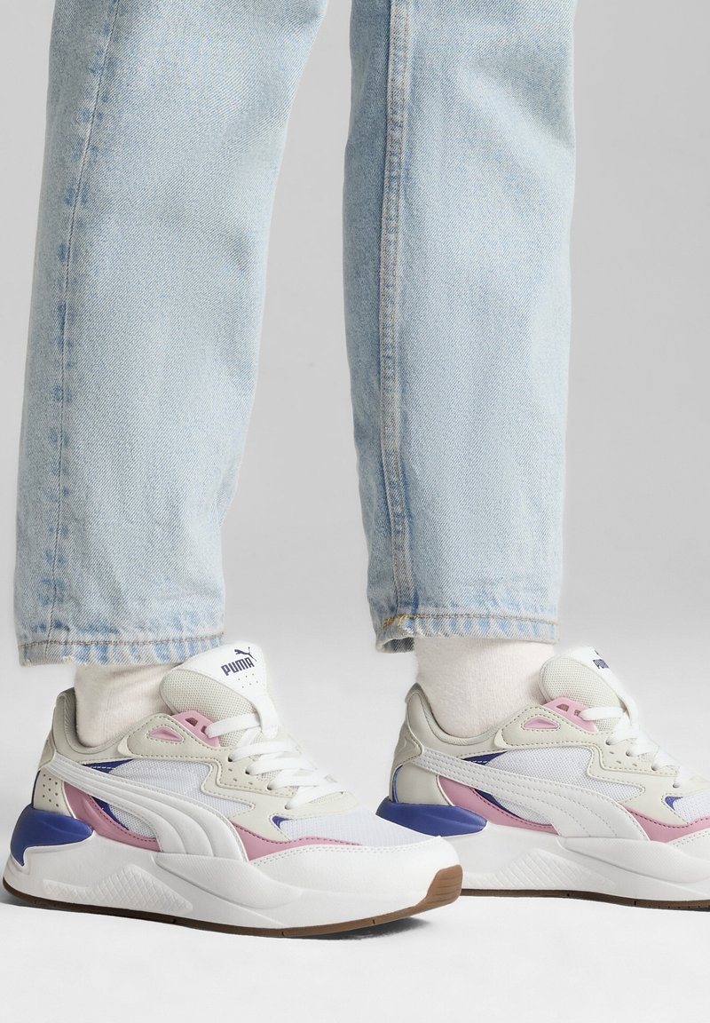 Weiße und graue Sneakers mit rosa und lila Akzenten, die eine strukturierte Sohle, auffällige Schnürsenkel und ein Logo auf der Zunge aufweisen. Getragen mit hellblauen Jeans.