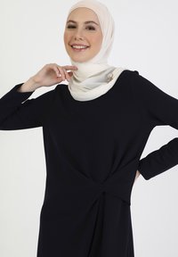 Dark navy dress with a twist design at the waist, featuring long sleeves and a round neckline. Accompanied by a light cream hijab with bead accents.