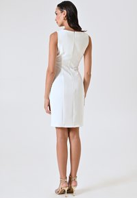 Robe blanche sans manches en tissu lisse, longueur mi-cuisse, dotée d'une fermeture éclair cachée dans le dos et d'une silhouette ajustée. Talons dorés à lanières.