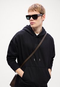 Zwarte hoodie met een voorzak, trekkoord halslijn en lange mouwen. Bruin schoudertasje strap kruist over de borst. Donkere zonnebril.