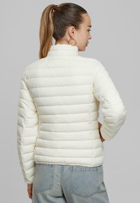 Veste blanche en duvet avec un design matelassé vertical et un col haut, associée à un jean denim bleu clair. La veste présente une texture lisse et brillante.