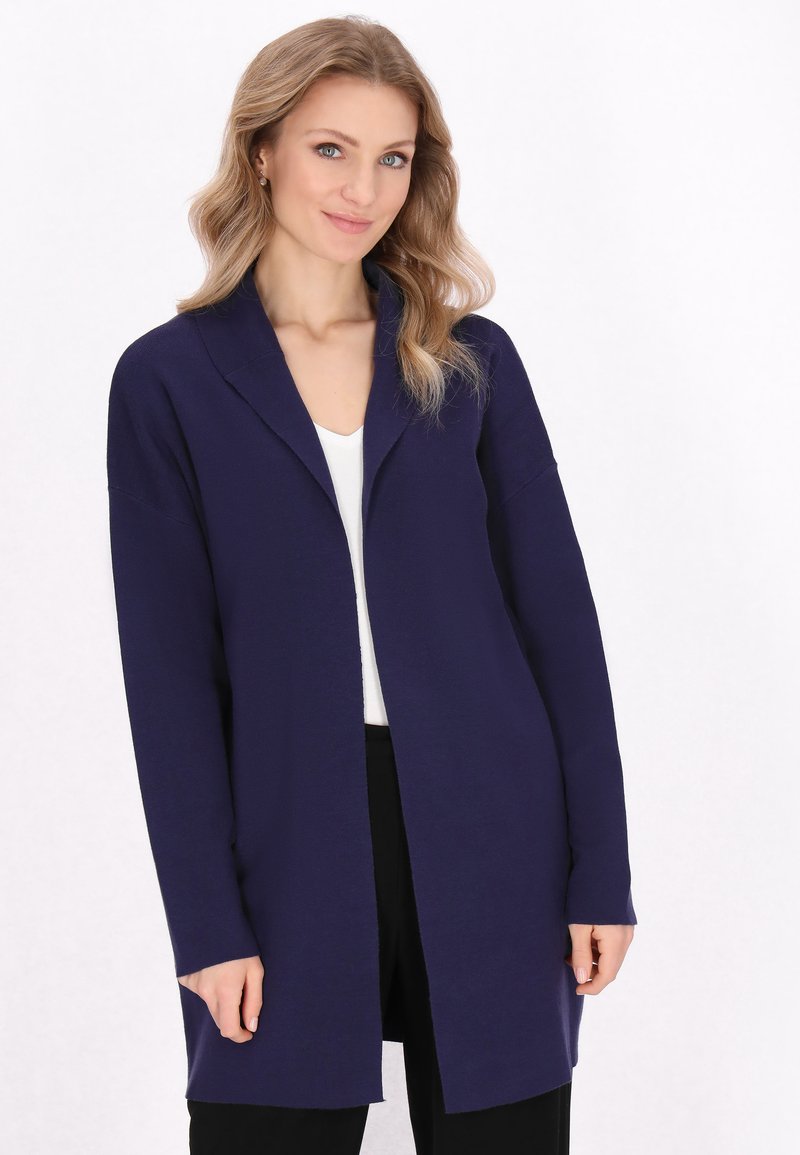 Cardigan lungo con apertura frontale in blu navy, realizzato in tessuto morbido. Presenta un colletto a scialle e spalle scese. Indossato sopra una maglietta bianca.