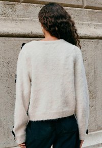 Cremfarbener, kuscheliger Pullover mit langen Ärmeln, der schwarze Bandakzente an den Bündchen hat. Strukturierter Stoff, lässige Passform und cropped Länge.