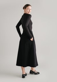 Czarna bluzka z długim rękawem zestawiona z ciemną, teksturowaną spódnicą sięgającą do podłogi. Modelka nosi czarne, pasekowe buty na niskim obcasie.