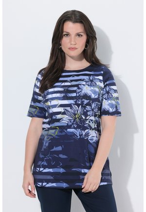 Frau mit langem dunklem Haar, die ein blaues, geblümtes und gestreiftes Kurzarmhemd trägt und vor einem schlichten hellen Hintergrund steht.