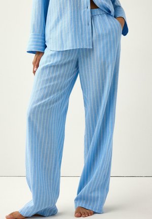 Persona che indossa pantaloni larghi azzurri a righe sottili e camicia abbinata, in piedi a piedi nudi con una mano in tasca su sfondo uniforme.